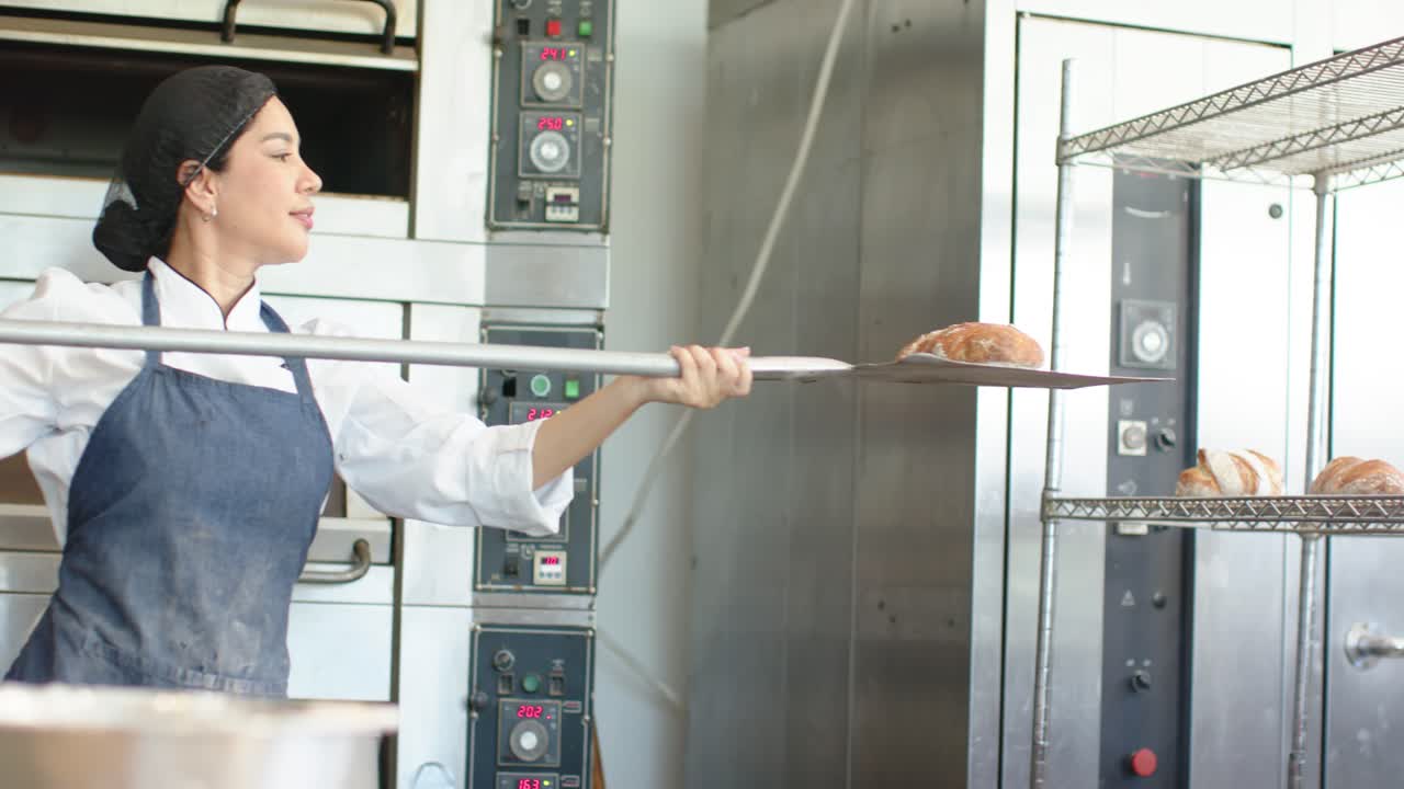 Una fornaia sente il timer del forno, rimuove le pagnotte con la pala e le mette su griglie di raffreddamento, indossando una giacca da chef.