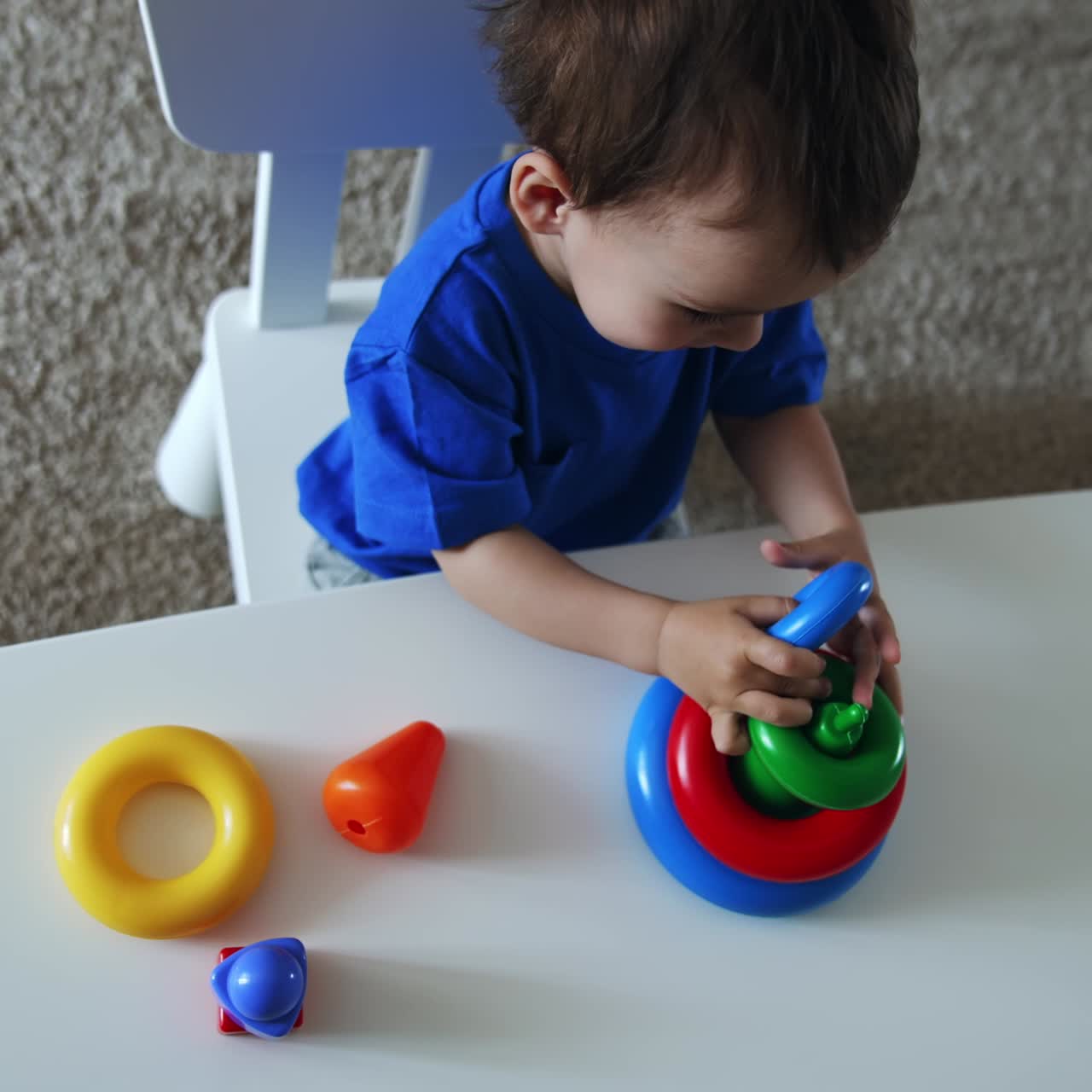 Dark-haired Caucasian child assembling toy pyramid. Charming kid plays sitting at desk indoors. Top view