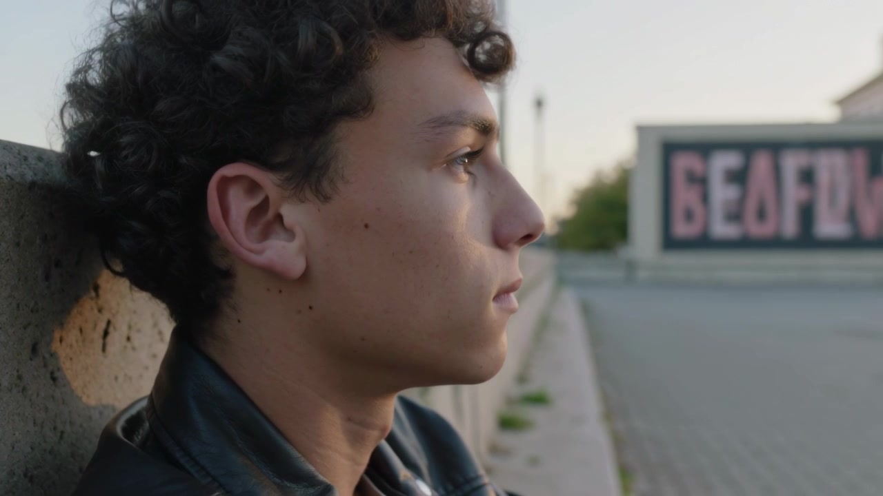 Adolescente con giacca di pelle appoggiato a un muro che si gira e guarda un cartellone rosa, con la telecamera che si allontana, spazio per il testo.