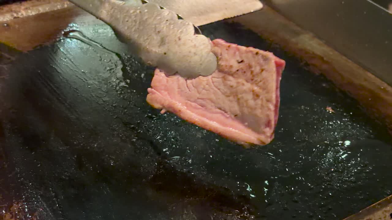 Cook flips thick wagyu steak on flat grill using tongs, under warm, indoor lighting