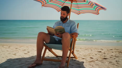 man reading a book on a beach under an umbrella