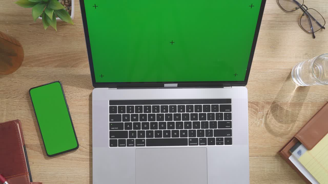 Top down view of a laptop computer with mock up green screen chromakey display on a wooden office desk next to notebook with pens, glasses, and a glass of water. Slow zoom out, close up