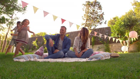 Children-Run-Around-Parents-As-They-Sit-On-Blanket-In-Garden