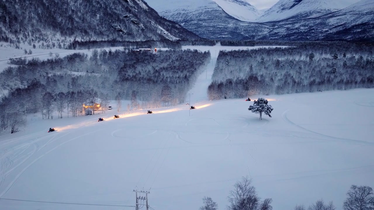 Motoslitte con i fari accesi procedono in fila su un paesaggio invernale innevato. Riprese da drone.