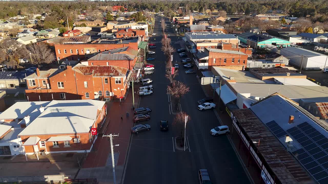 Drone footage captures vehicles moving along a sunlit main street lined with brick buildings in a quiet Australian town, with steady forward camera motion