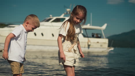 attentive brother and sister stand knee deep in sea with warm temperature water. luxurious yacht provides stunning background all bathed in warm glow of sun