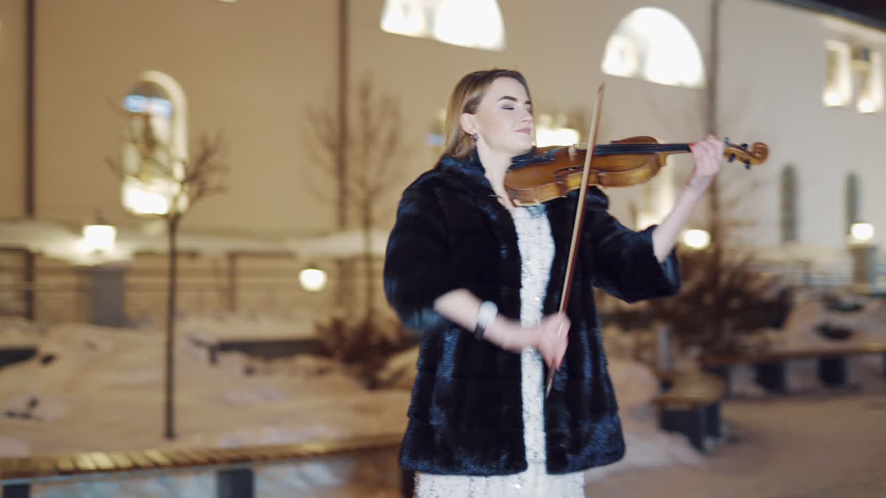 An attractive girl is standing in the square in a long white dress with sequins and playing the violin on the background of the monument and the buildings behind her in the evening. Blurred background.
