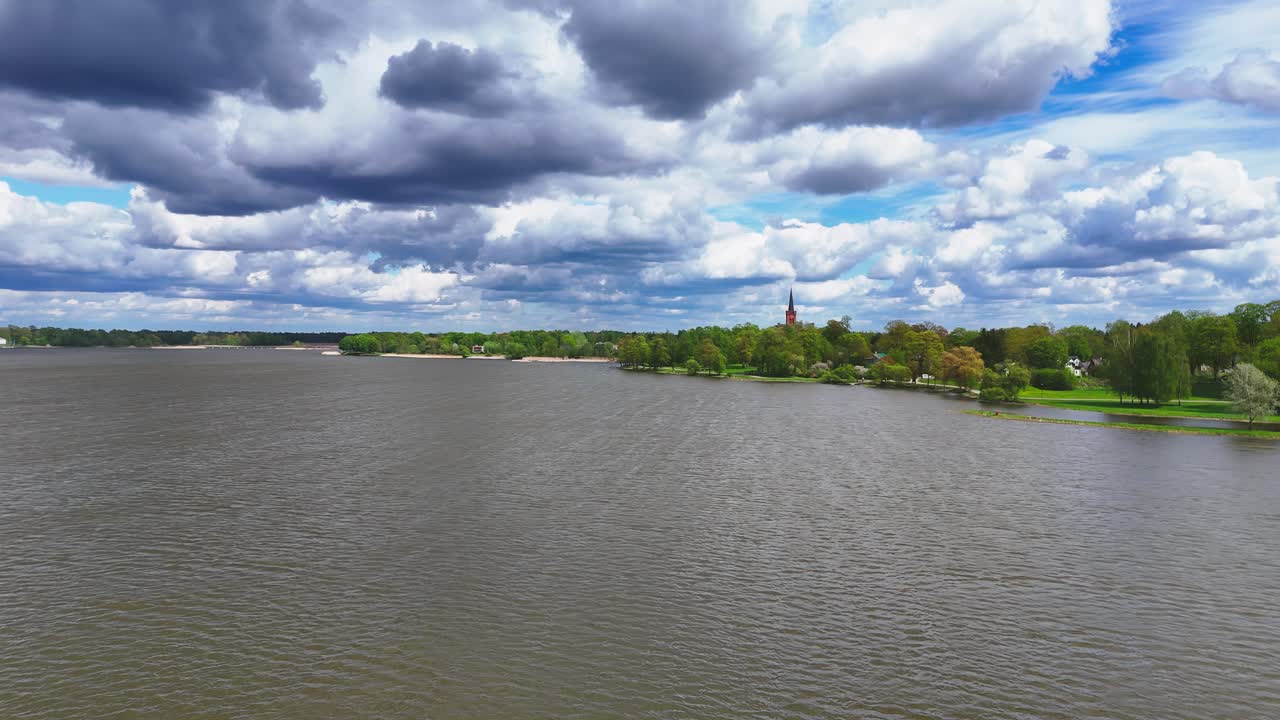 elevated, wide-angle view of Lithuania's Širvėna Lake in Biržai during a partly cloudy spring day, showcasing the grey-brown water, distant green shoreline with a red church spire