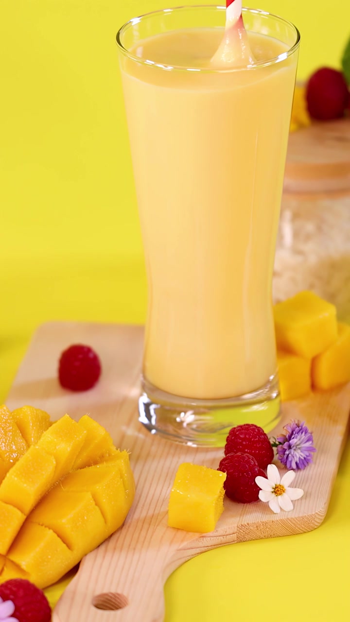 Smoothie al mango versato in un bicchiere, circondato da cubetti di mango, lamponi, fiori, sfondo giallo brillante