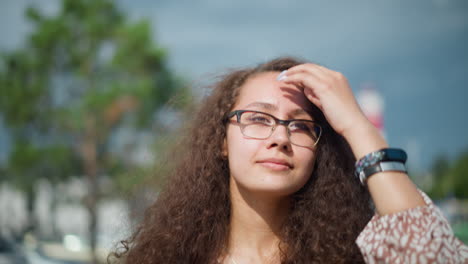 mujer con cabello rizado y gafas caminando al aire libre con una cálida sonrisa, ajustando el cabello hacia atrás bajo la luz del sol, con edificios borrosos y vegetación en el fondo