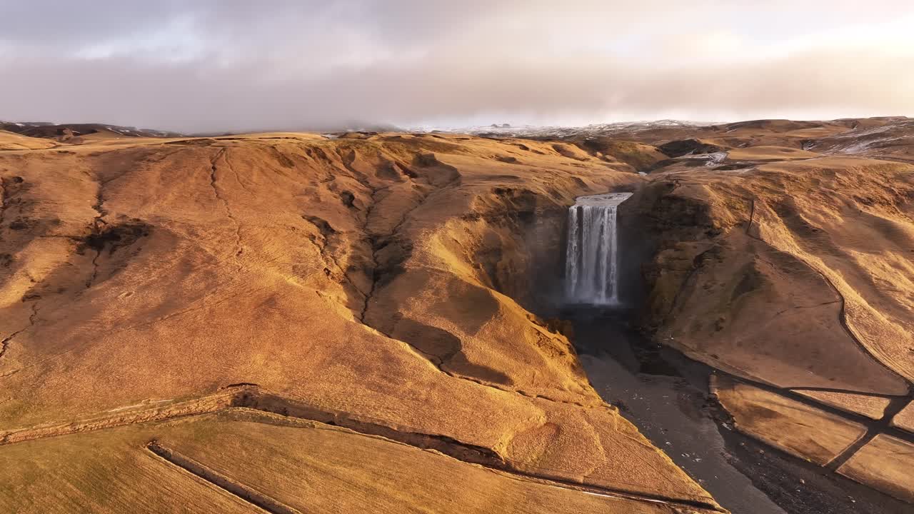 Aerial view of Skógafoss waterfall in Skógar, Iceland, with golden winter terrain, winding paths, and Skógá river cascading dramatically from the cliffs near Eyjafjallajökull volcano.
