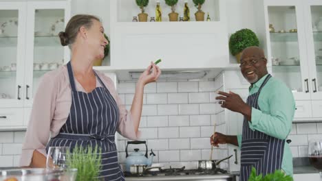 smiling senior diverse couple wearing blue aprons and cooking in kitchen