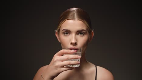 Serious-woman-takes-a-sip-out-of-a-transparent-water-glass,-black-background