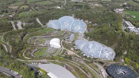 sweeping aerial of eden project geodesic biomes gleaming within reclaimed quarry amid terraced gardens and forest, showcasing eco-tourism, biodiversity and sustainable innovation