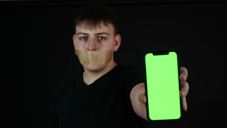 cyber bullying concept, young man with tape over mouth holding smartphone with green screen