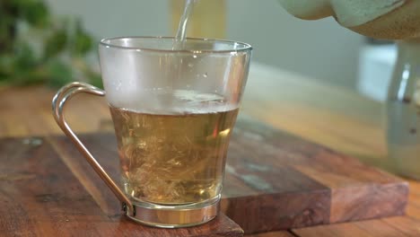 making herbal tea in a green teapot on a wood table in a light and airy room with green plants in the background and a glass teacup