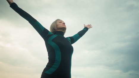 woman standing outdoors under cloudy sky practicing yoga, lifting her hands up and bringing them down slowly, wearing green and black fitness outfit, she focuses on calm movement