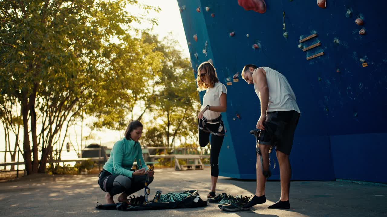 dos chicas en uniformes deportivos y un tipo en una camiseta blanca se pusieron equipo especial y seguro antes de escalar en una pared de escalada. un grupo de escaladores se calientan antes de escalada en la pared de ascenso en un soleado día de verano