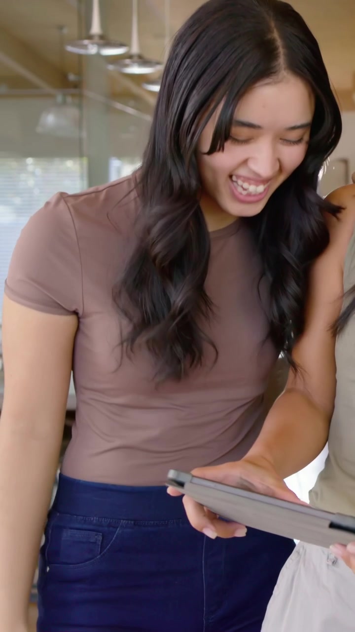 Video verticale: Donna con top marrone che tocca lo schermo, chinandosi mentre un'amica tiene un tablet al bar, entrambe ridono.