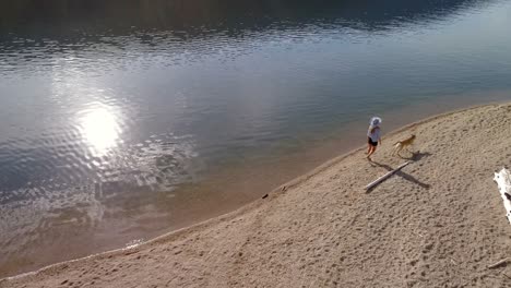 Woman-with-her-pet-dog-walking-near-river-coast-on-a-sunny-day-4k