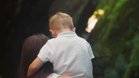 young mother and preschooler boy stand on path resting together in nature reserve backside view. woman holds child in arms talking calmly closeup