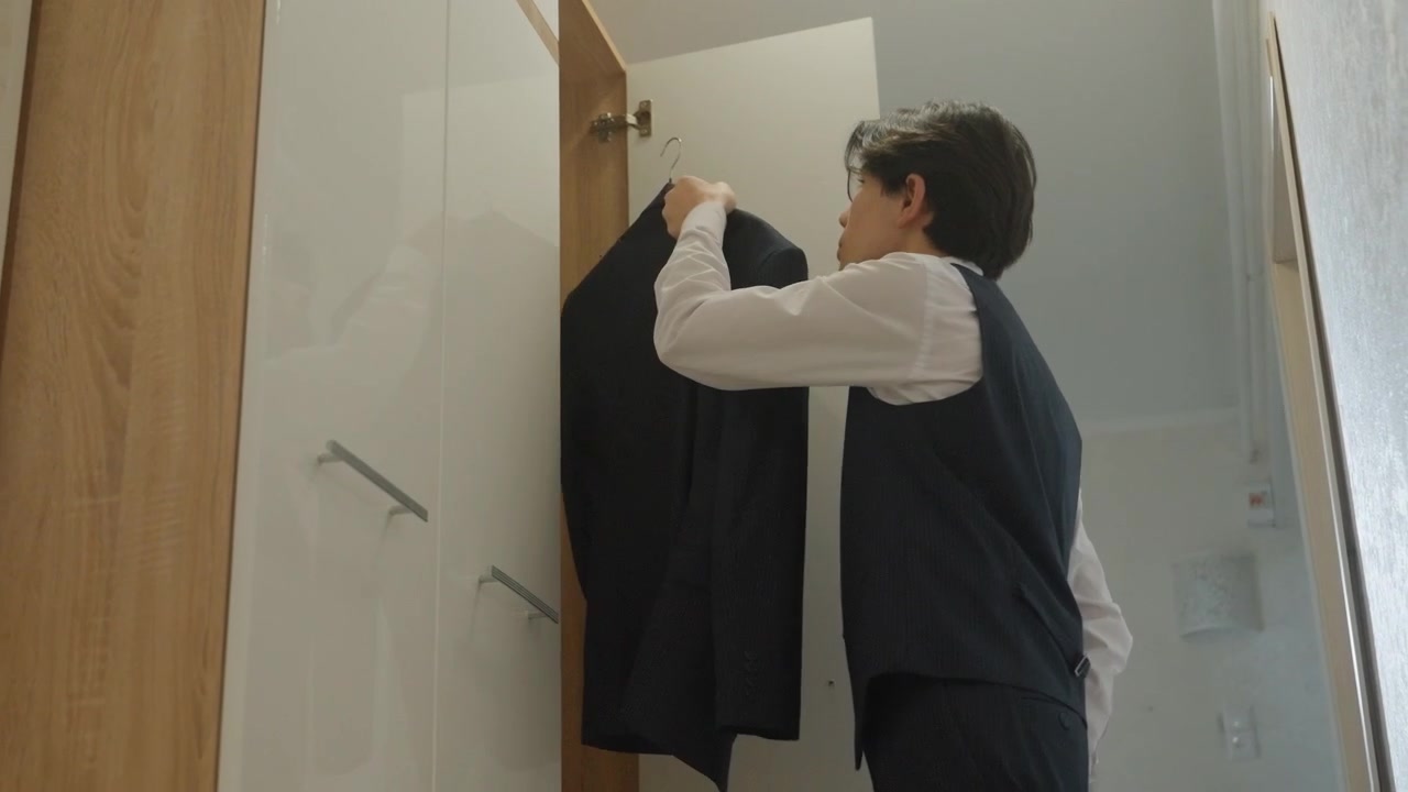 Giovane uomo nel corridoio che controlla l'armadio, tiene in mano la giacca, routine mattutina contemplativa con gilet e cravatta, luce naturale soffusa, interno di appartamento minimalista, movimenti lenti e deliberati, quiete.