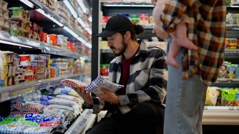 Brunette-guy-consults-with-his-wife-about-the-choice-of-dairy-products-during-family-shopping-in-the-supermarket.-A-brunette-girl-together-with-her-husband-and-infant-choose-goods-in-a-supermarket