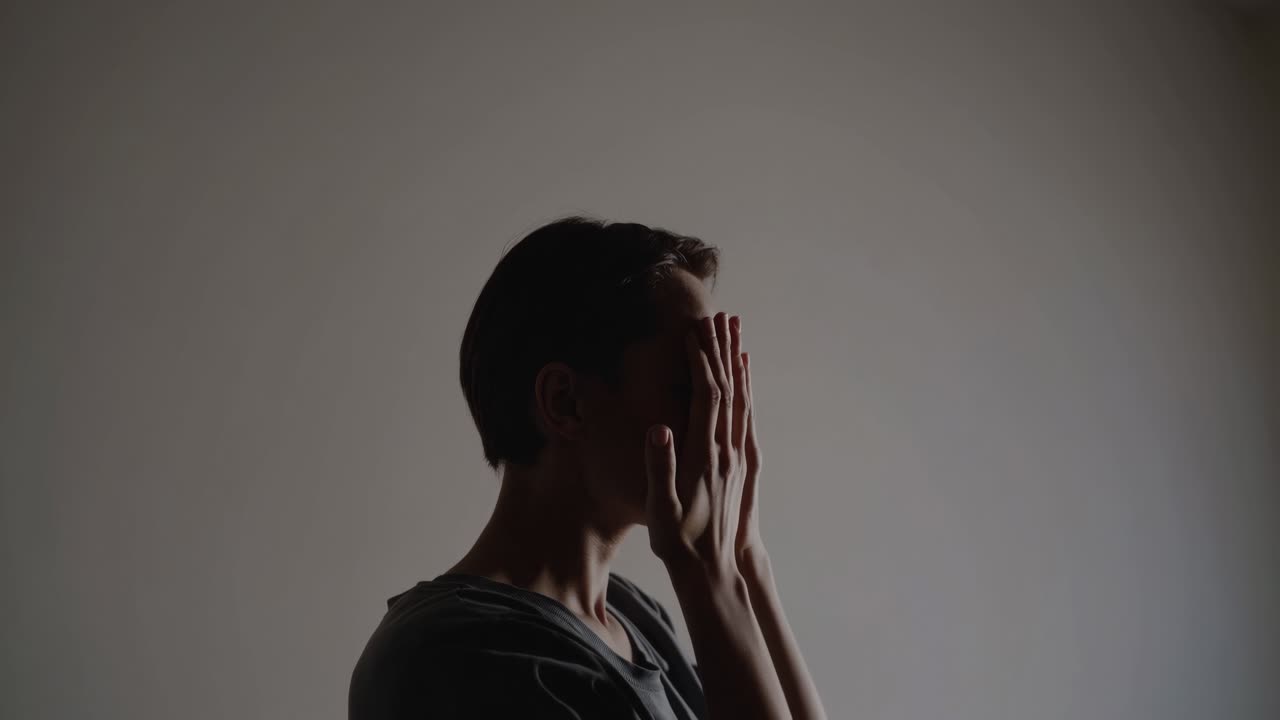 Uma filmagem de vídeo sombria, em ângulo lateral, de uma pessoa cobrindo o rosto com as mãos, contra um fundo liso.