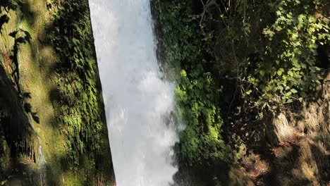tomando una panorámica de una cascada de agua cristalina rodeada por un acantilado cubierto de musgo para revelar que se derrama desde el borde de un acantilado desde la base de una cascada donde desemboca en un charco de agua.