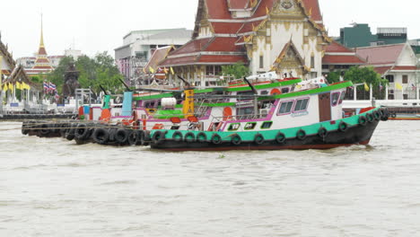 colorful tugboats moving simultaneously in chao phraya river, passing by a local buddhist temple in bangkok, thailand.