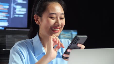 close up of asian female programmer using smartphone while writing code by a laptop using multiple monitors showing database on terminal window desktops in the office