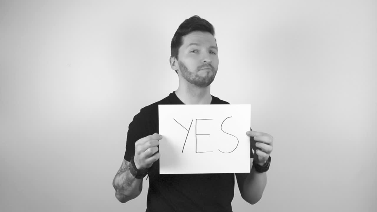 Grayscale footage of a tattooed man lifting up a white paper or a sing that says YES in capital letter and he is happy and confident while nodding when looking straight ahead into the camera.