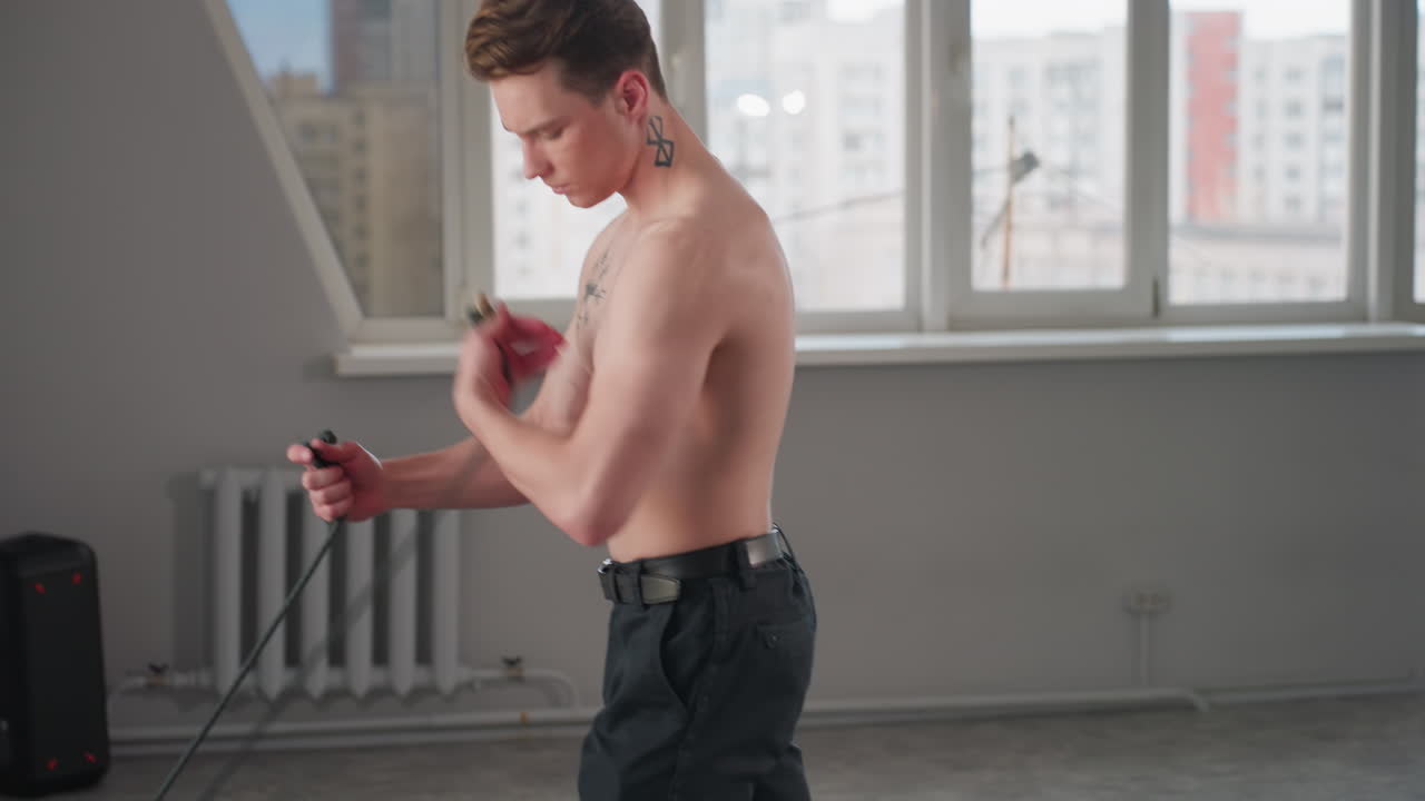 shirtless young man stands in bright loft studio, grips heavy black rope with both hands, braces legs and pulls rope across floor in powerful workout move