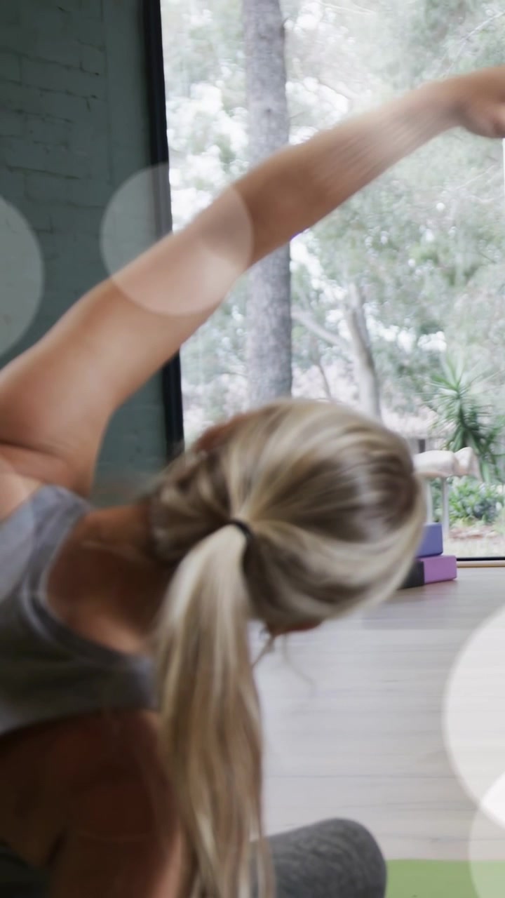 Vídeo vertical: Mulher levantando o braço direito, alongando-se lateralmente para fitness perto da janela com tapete