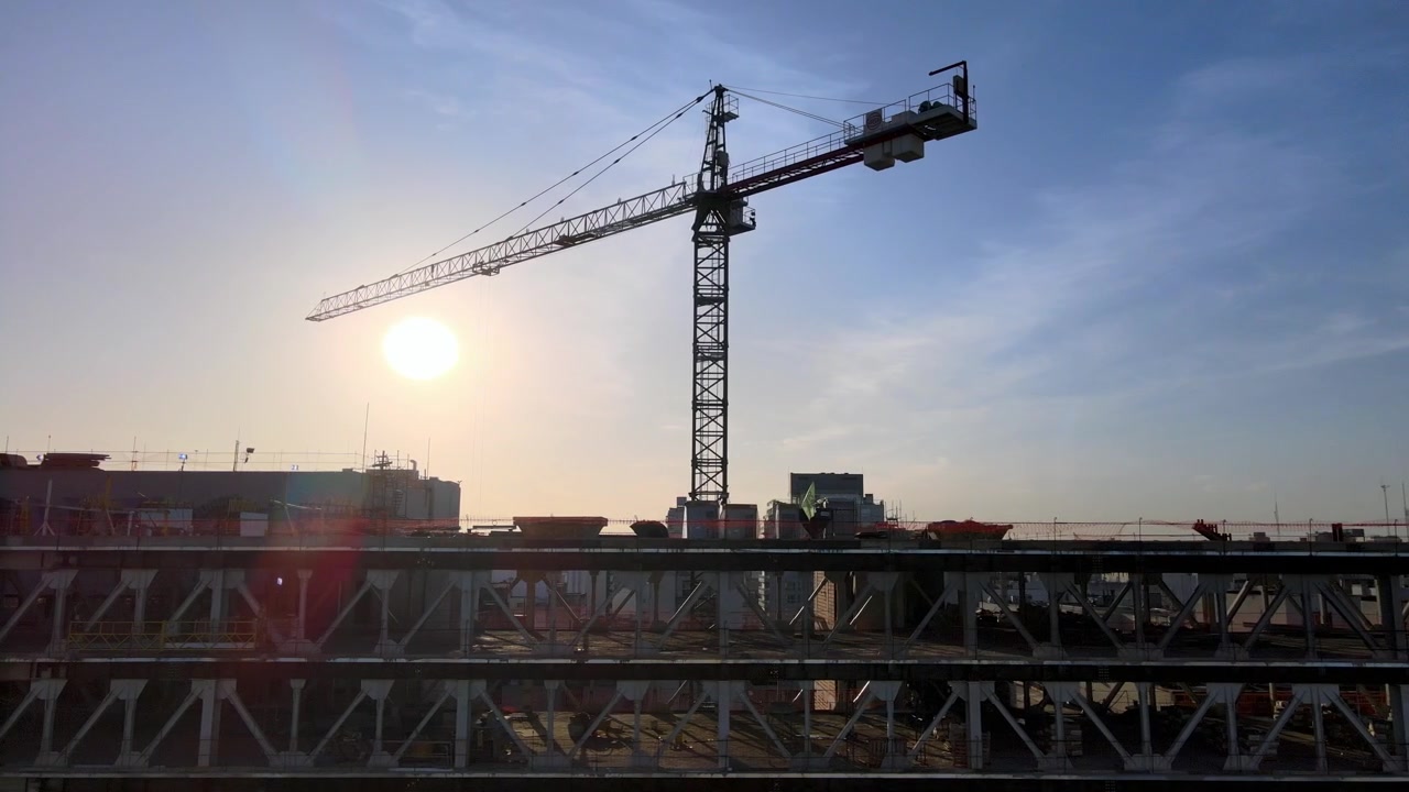Panning drone shot of construction crane on site. Camera follows building under construction. Crane towers up in blue sky with sun behind. 23 sec.