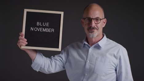 ritratto in studio di un uomo maturo che tiene in mano un cartello che legge "blue november" che promuove la consapevolezza della salute degli uomini e del cancro 1