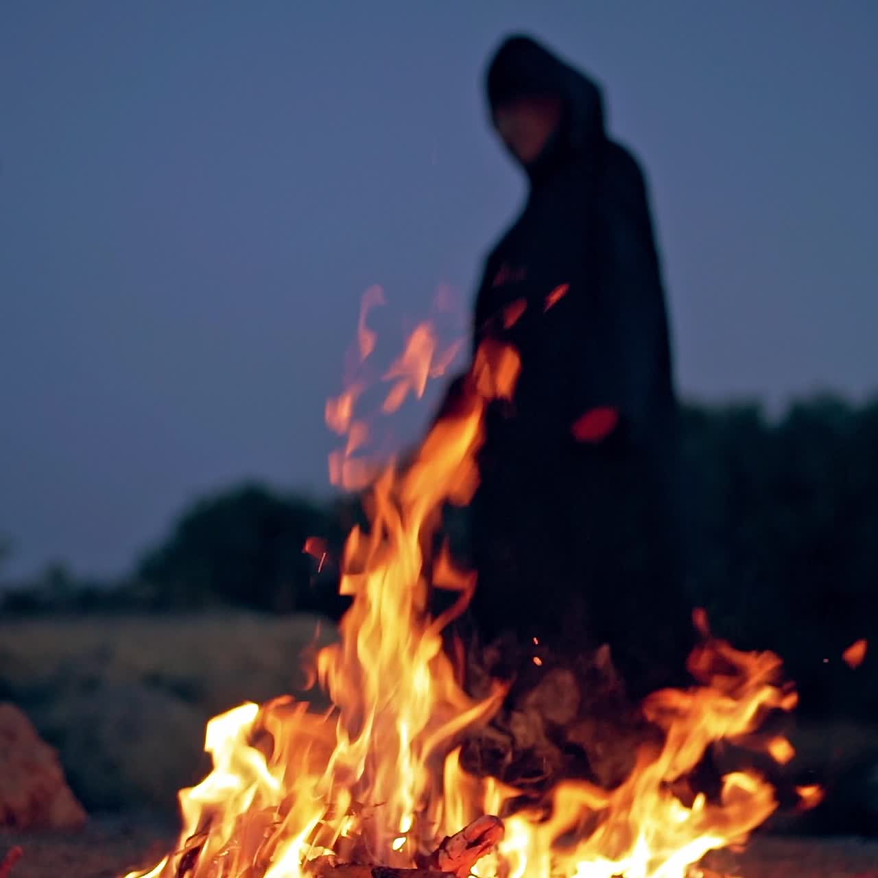Bright fire flame and black witch behind. Spooky figure in black cloak standing near the burning fire at night. Halloween concept.