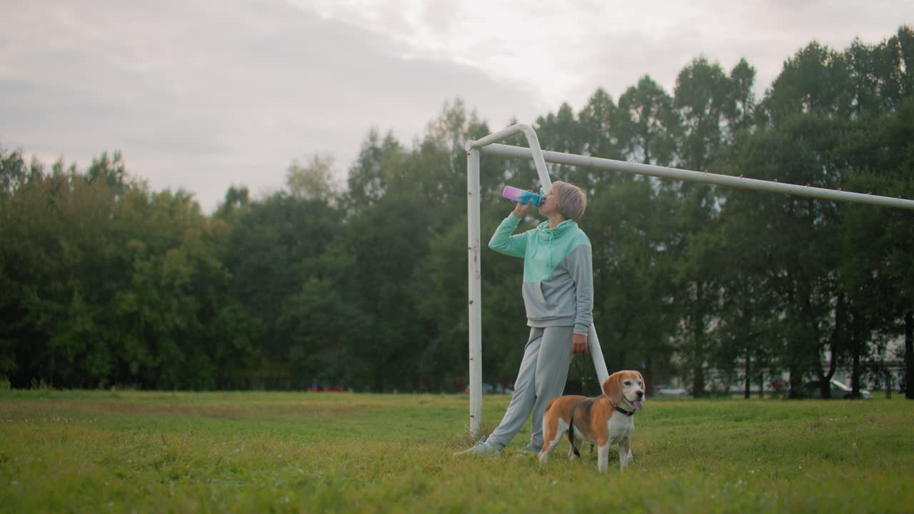 Sport woman dressed in gray and mint tracksuit resting on goalpost while drinking water from colorful bottle accompanied by her athletic beagle dog on grassy field during outdoor exercise session