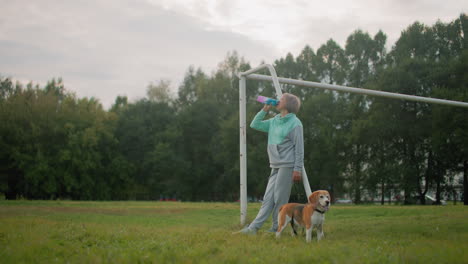 sport woman dressed in gray and mint tracksuit resting on goalpost while drinking water from colorful bottle accompanied by her athletic beagle dog on grassy field during outdoor exercise session