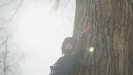 a young boy leans against a large tree trunk with sunlight shining directly on him, his arms are outstretched, embracing the moment, he is dressed in a shiny black jacket, with trees around