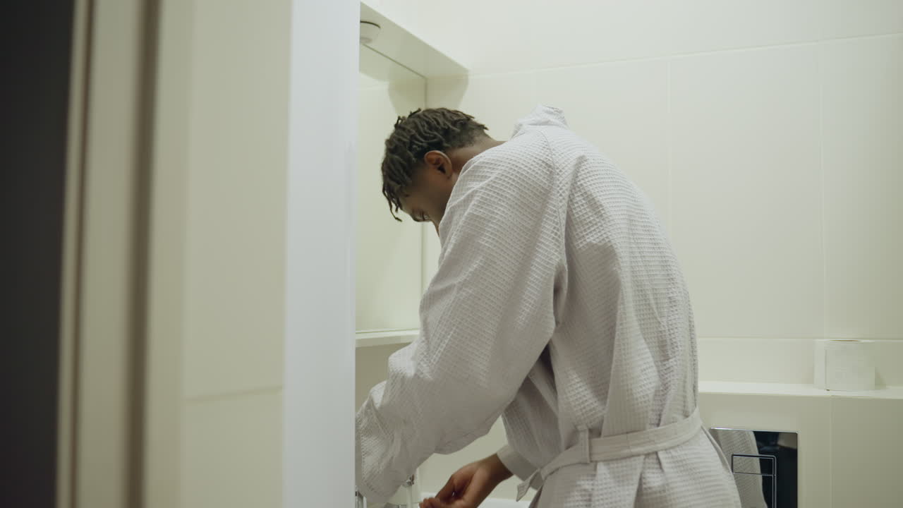 Black youth in robe standing at bathroom sink, washing hands, relaxed indoor setting, hygiene routine, clean modern bathroom with soft lighting, daily self-care, morning preparation