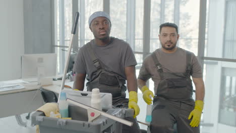 and arabic cleaning man wearing gloves posing with cleaning cart and looking at the camera inside an office