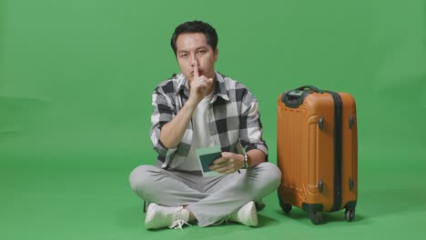 full body of asian male traveler with luggage and passport looking at camera and making shh gesture while sitting in the green screen background studio