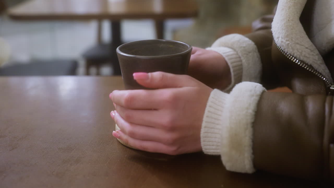 Close-up of lady hand in brown shearling jacket gently placing cup of coffee on table in cozy cafe. Calm, inviting atmosphere with blurred background, enhancing the peaceful moment