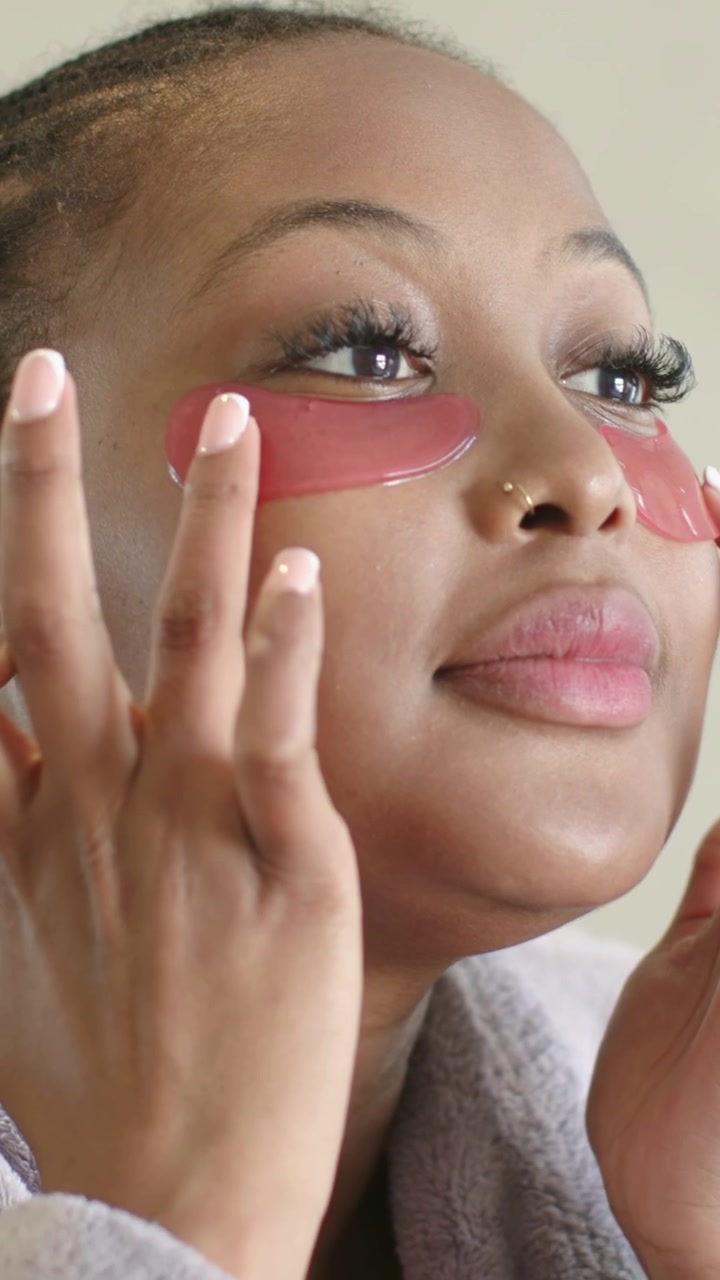 Vertical video: Bringing hands, woman applying pink undereye patches at home in robe to hydrate