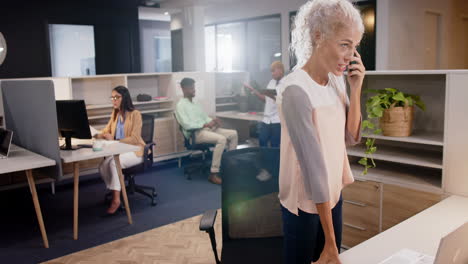 talking on phone, woman standing in modern office with colleagues working
