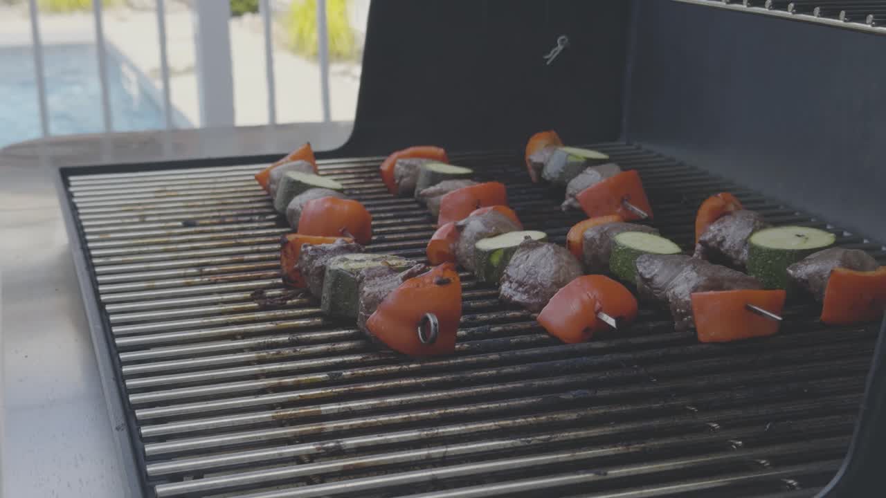 Side angle of Beef, zucchini, and red bell pepper skewers cooking on a stainless steel BBQ grill. Stainless steel tongs adjust the skewers over the heat