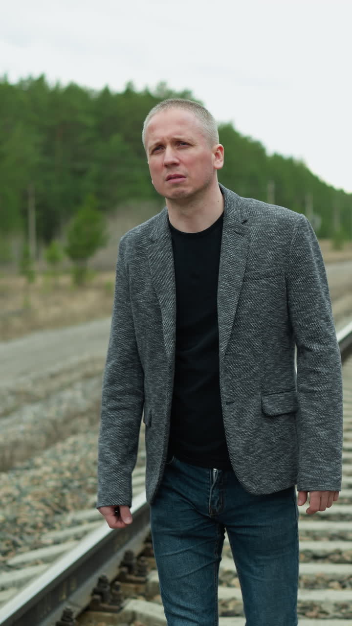 A close-up view of a middle-aged man wearing a grey suit jacket and black shirt, walking along railway tracks in a forested area