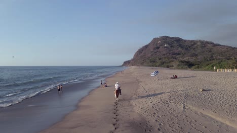 aerial follows cowboy riding horse on sand beach as tourist enjoy sea
