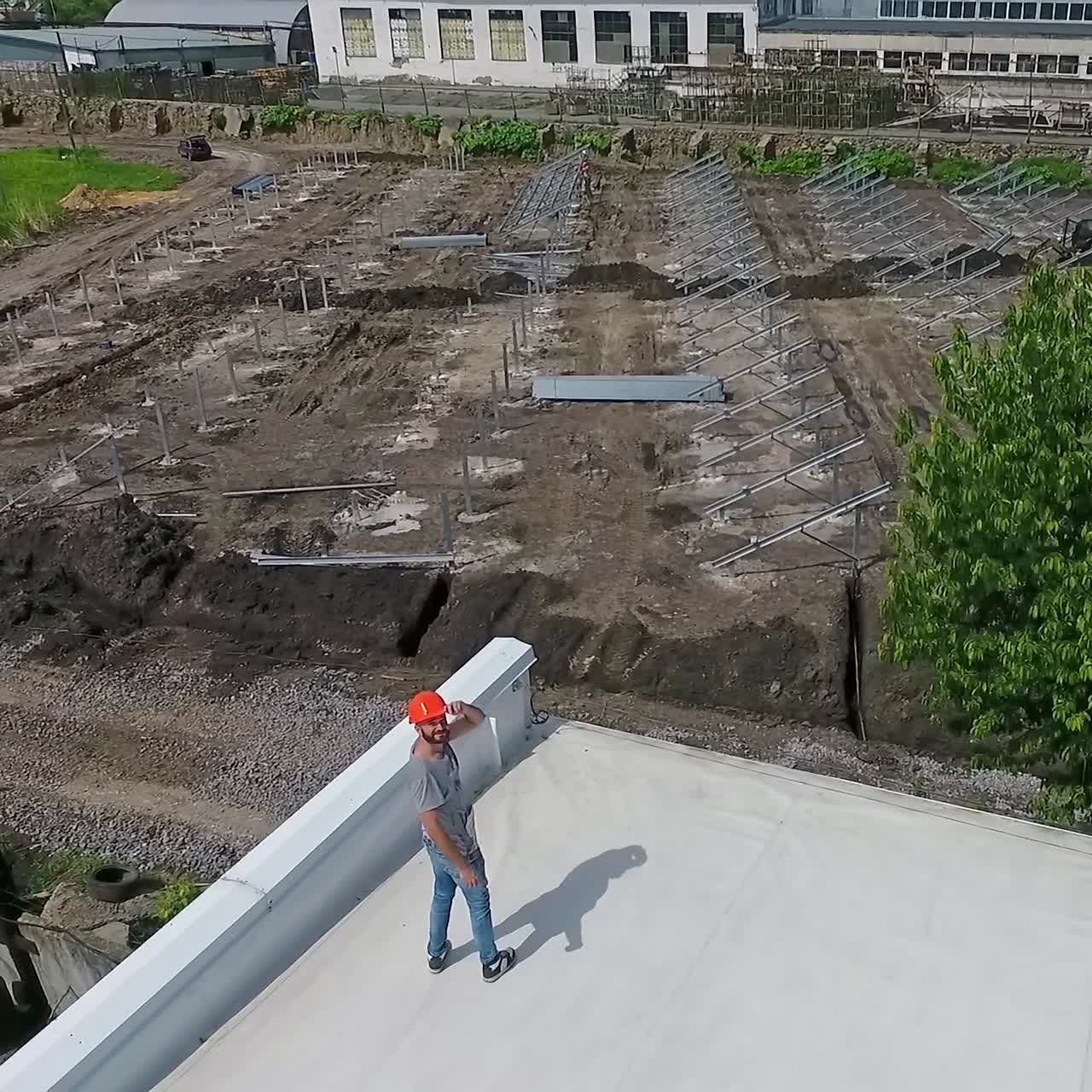 Technician in orange hard hat on a roof in summer. Installer looking on the solar farm construction from a rooftop of a building. Aerial view.